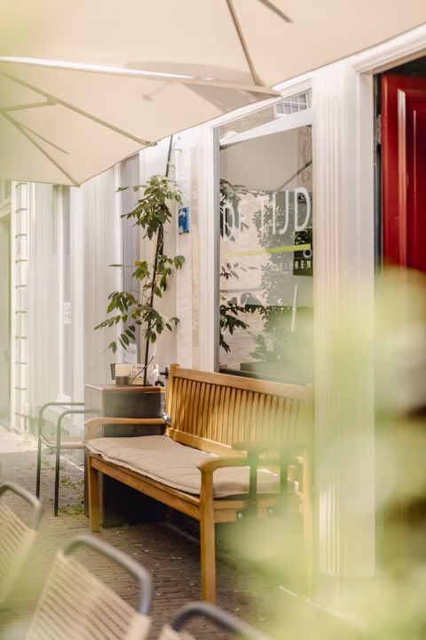 the cafe terrace shot through some plants with wooden furniture and a the red front door in the background